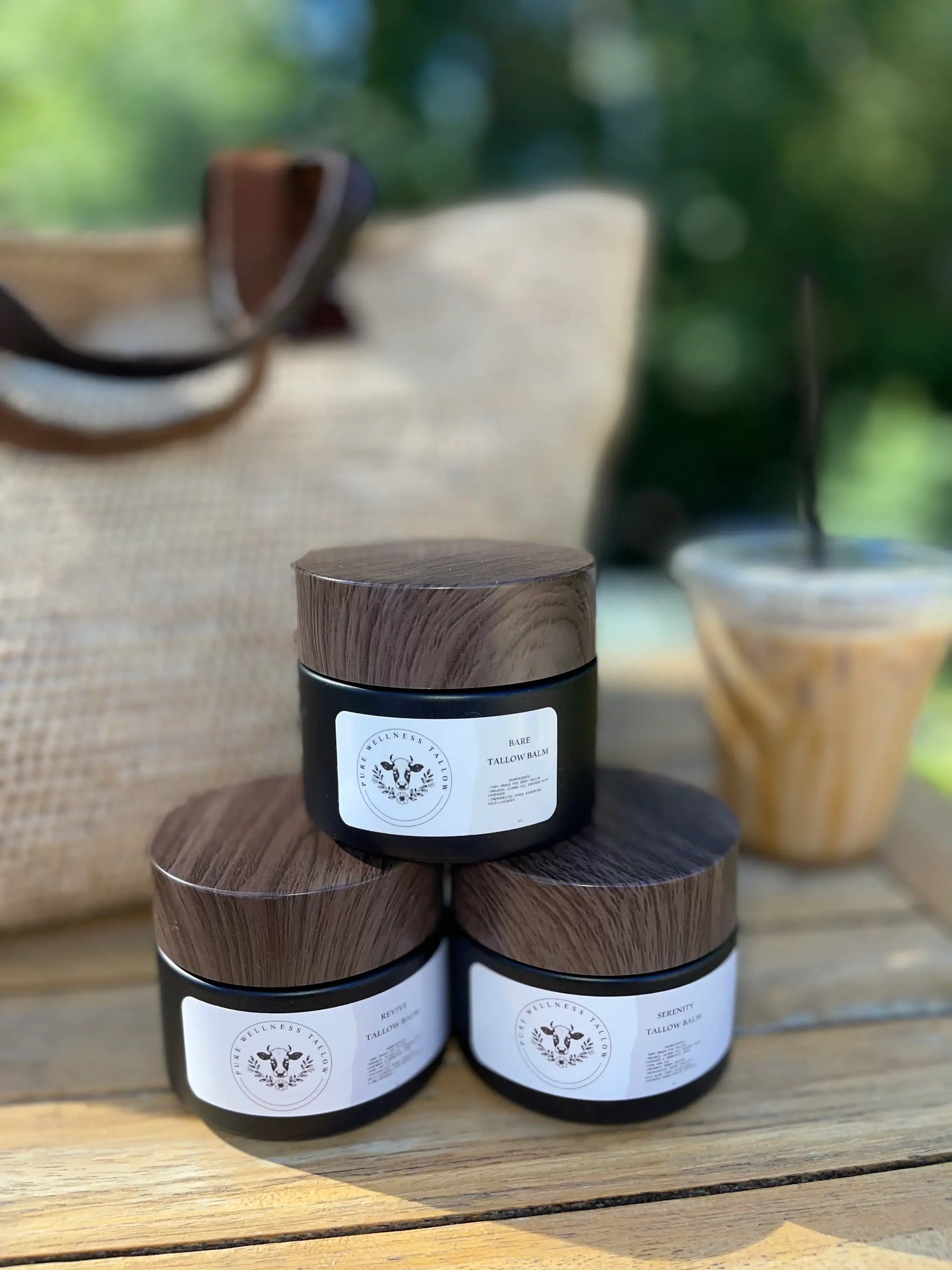 Three natural skincare jars on a wooden table with a rustic bag and iced coffee in the background.